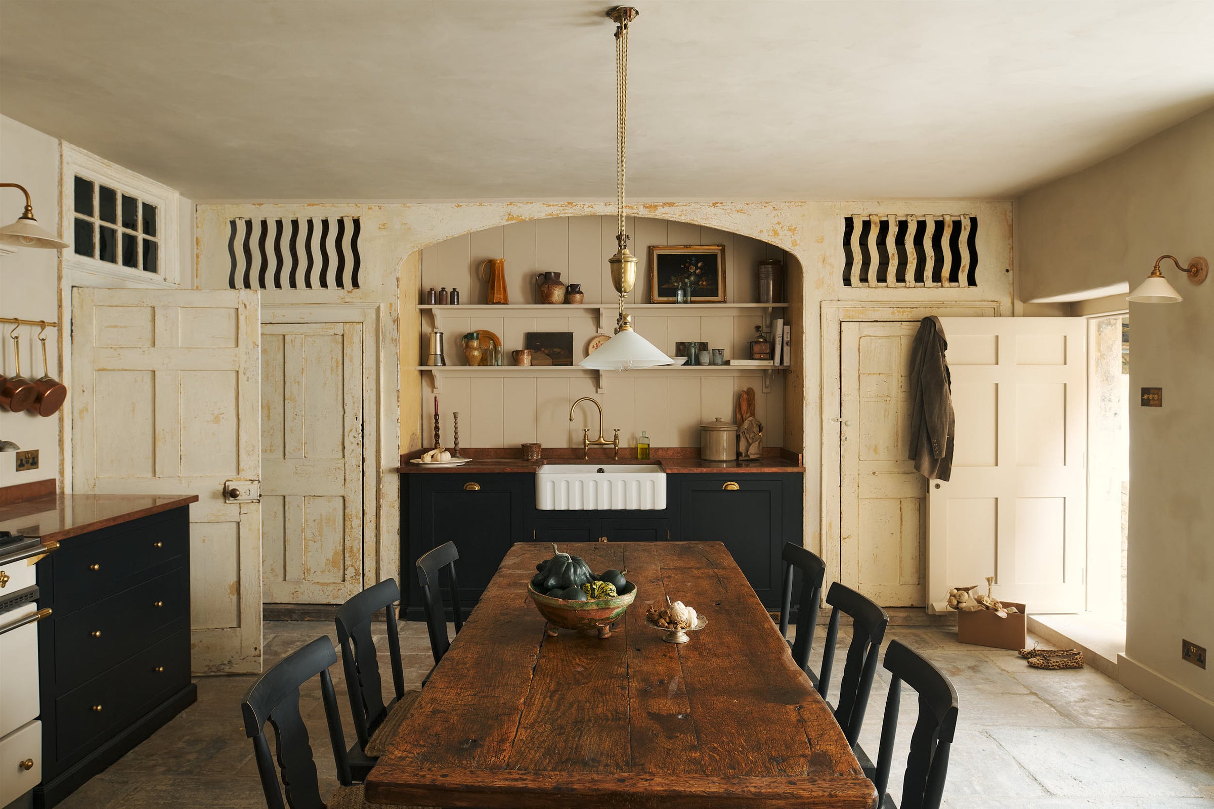A rustic basement kitchen with sandy-coloured walls and ceiling, original Georgian detailing, cream doors and panelling with chipped paintwork and a worn stone floor. An old banquet-style table sits in the middle of the room with rows of black wooden chairs on either side. At the far side of the roo