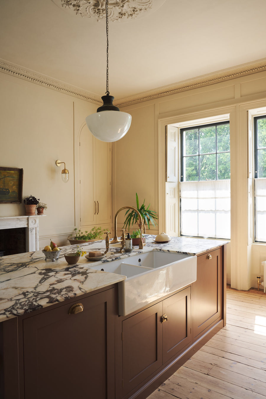 An oblique view of a bright, spacious, warm, and simple-yet-grand shaker kitchen featuring a large kitchen island with a Calacatta Viola worktop, and an aged brass Ionian tap. Perfectly decorated with beloved vintage furniture and decor made from different natural materials, it creates a scene of ab