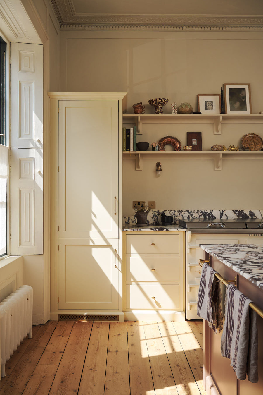 A quiet corner of a bright, spacious, warm, and simple-yet-grand shaker kitchen featuring a large kitchen island with a Calacatta Viola worktop, a cooker run, a fridge/freezer cupboard, and long shaker shelves. Perfectly decorated with beloved vintage furniture and decor made from different natural 