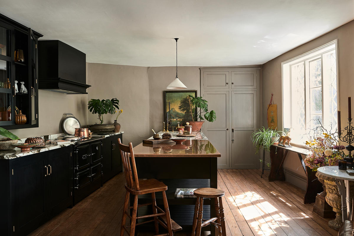 Bright light floods an understated Painter’s Black Shaker kitchen featuring an aged copper sink, aged brass taps, and a San Simone quartzite worktop—its pale white base accented with bold dark grey veins and subtle gold flecks. Grey walls frame the space, with a cheese plant and vintage oil painting