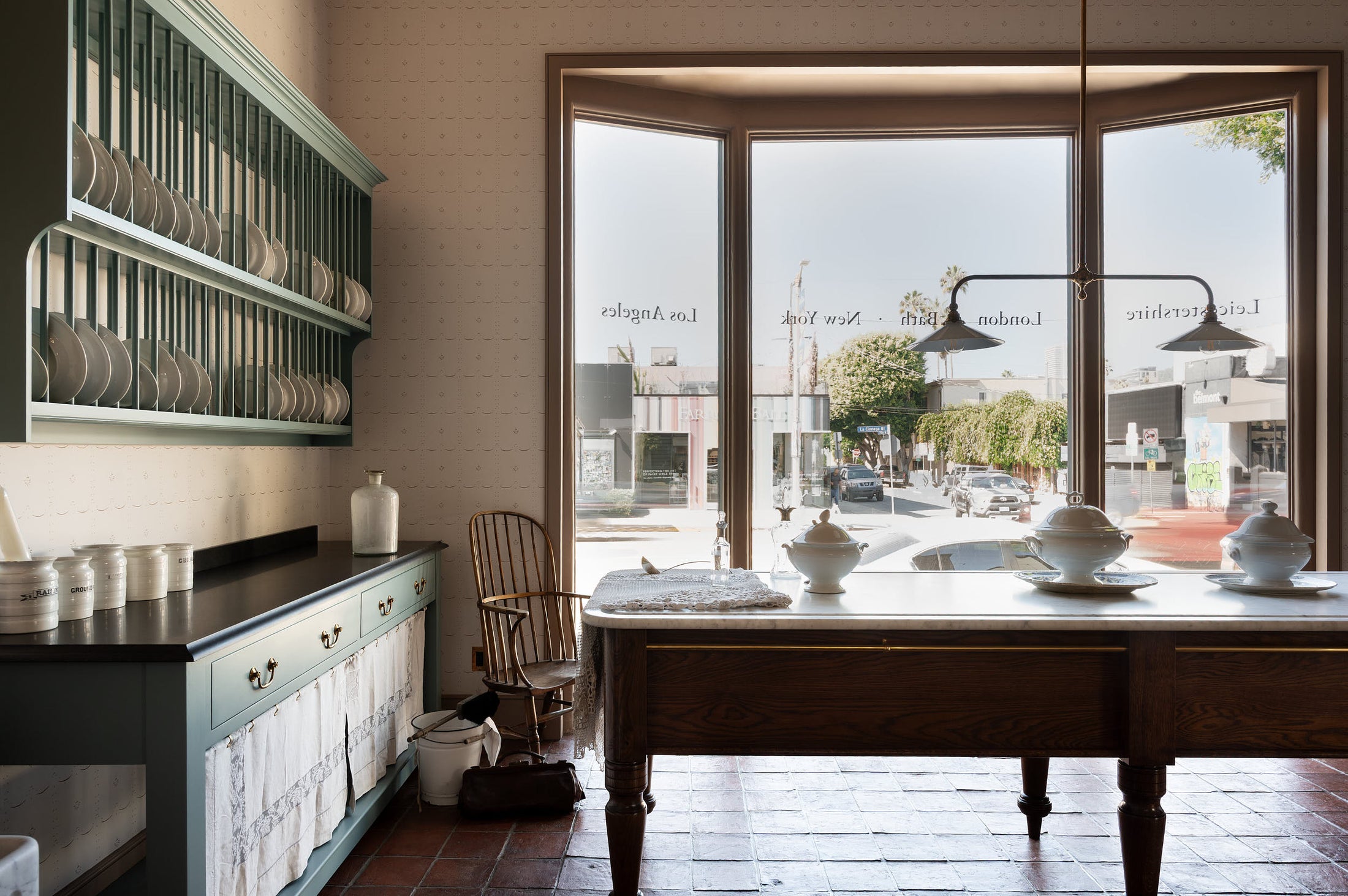 A grand and spacious Heirloom kitchen with Trinity blue open cabinets and plate racks, Heirloom Dairy Tables, an Heirloom Gaselier light reaching all the way to the top of the high ceiling and rustic Square Terracotta tiles. Beautifully decorated with a spectacular chandelier, artisan ceramics, and