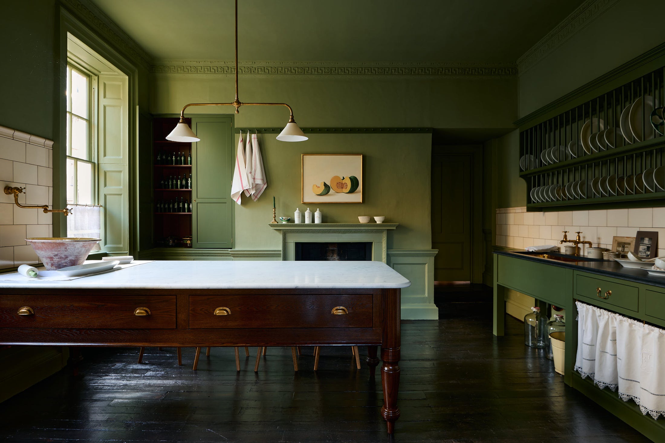 A quaint and stylish Wilkes green Heirloom kitchen features a sink cabinet with a café rail, white lace curtains, a polished brass sink, aged brass Mayan taps, a large plate rack, and a dairy table. Adorned with an aged brass pot filler, artisan ceramics, Heirloom gaselier lights, cream subway splas