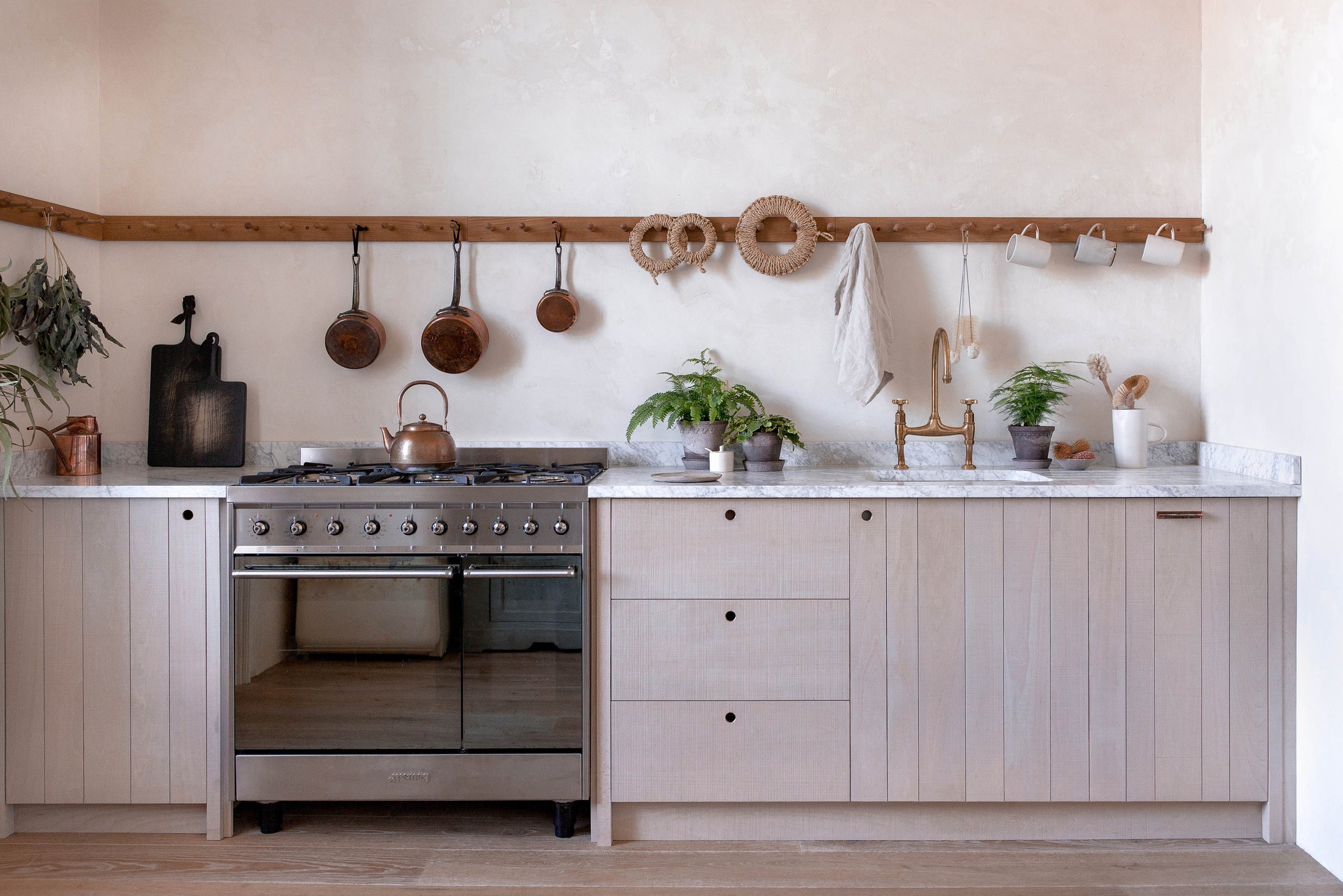 A light contemporary kitchen with creamy textured walls, rough-sawn beech slatted cupboard doors, an aged brass tap and a span of wooden pegs for hanging mugs, pans and utensils above a mottled white marble worktop. The styling is simple, Scandinavian and slightly rustic, with small potted ferns and