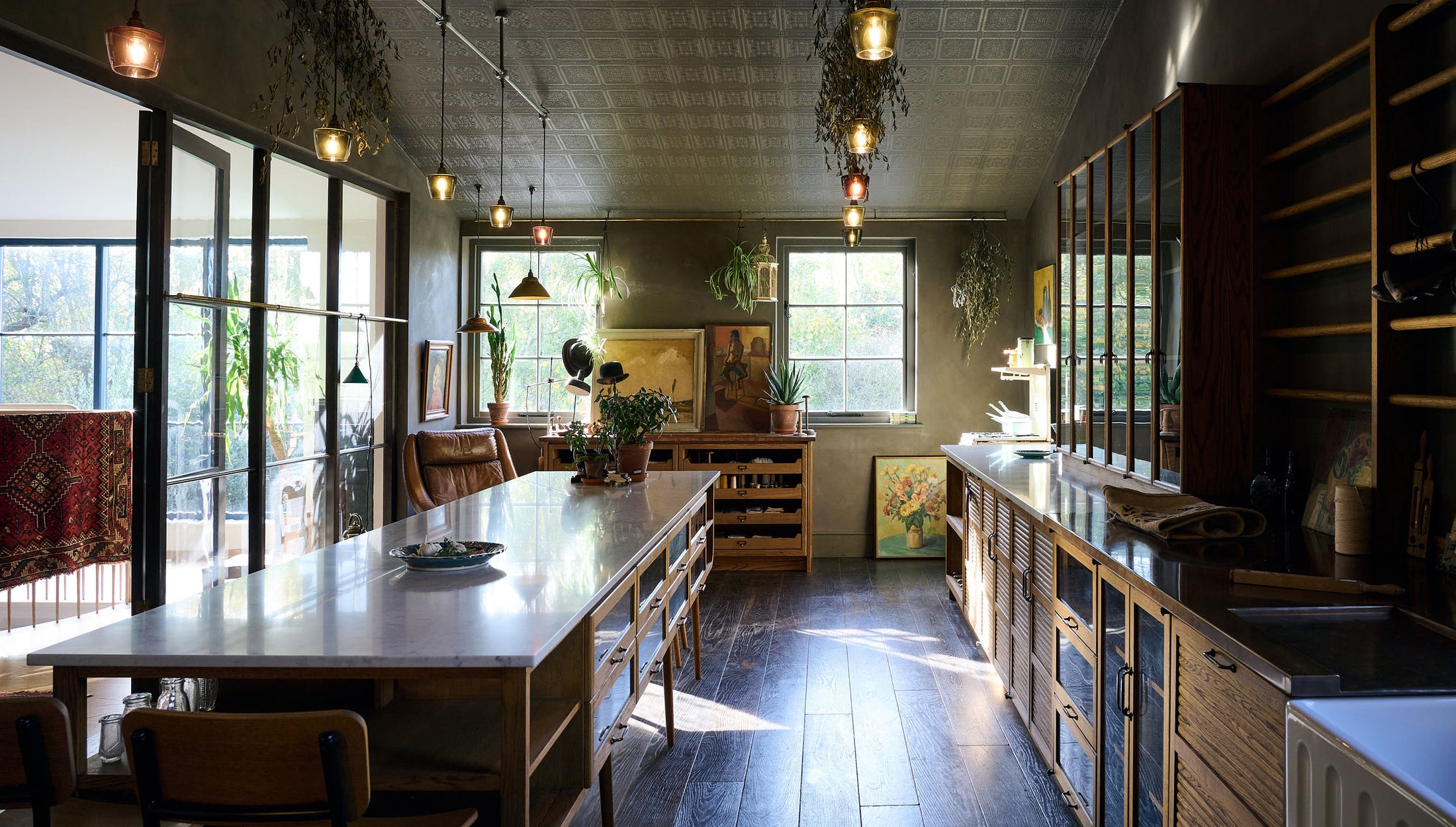 A warm and inviting Haberdasher’s kitchen features a long sink run with a Carrara marble and copper worktop, a double-fluted marble sink, a kitchen island, wall cupboards, and countertop cupboards. Adorned with luscious green hanging and potted plants, vintage items, artisan ceramics, illustrative t