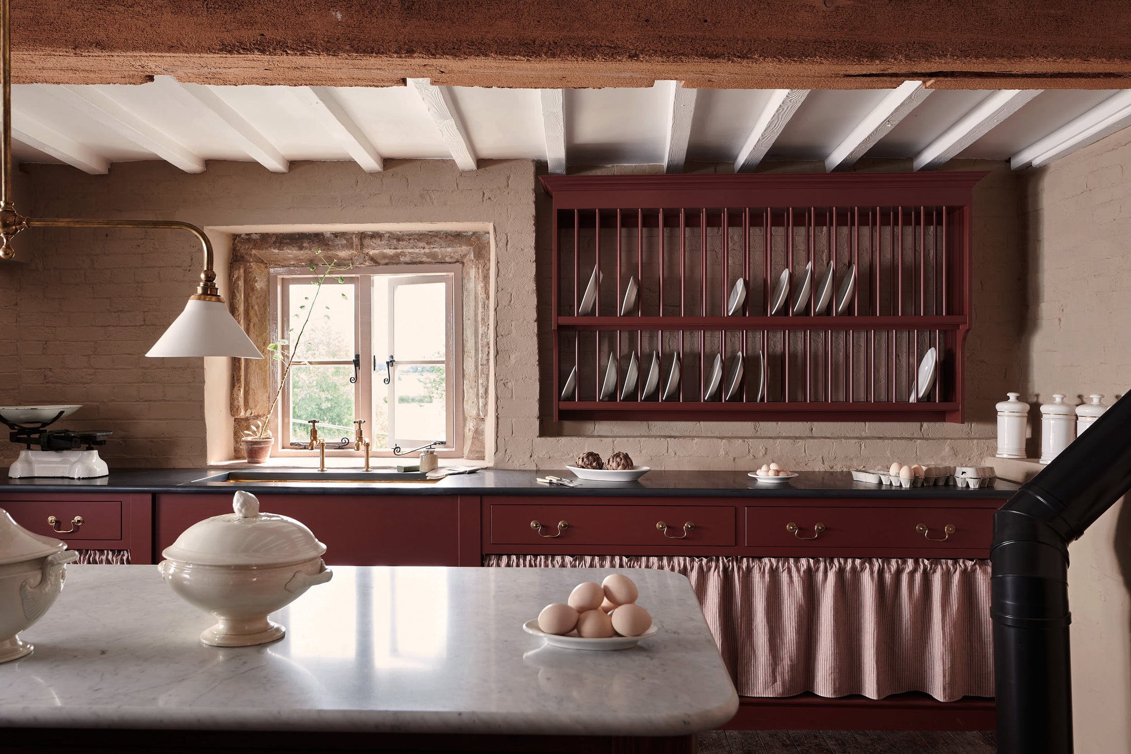 A refectory-red Heirloom kitchen’s sink cabinet reveals a polished brass sink, aged brass Mayan taps, café curtain rails, a red and white striped curtain, a plate rack, and an Heirloom dairy table. Beautifully decorated with Heirloom gaselier lights, a vintage scale, artisan ceramics, and a plate of