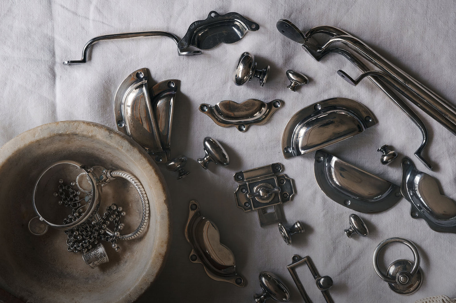 A collection of metal door knobs, handles and catches for cabinets in an Antique Silver finish, laid out on a white linen tablecloth.