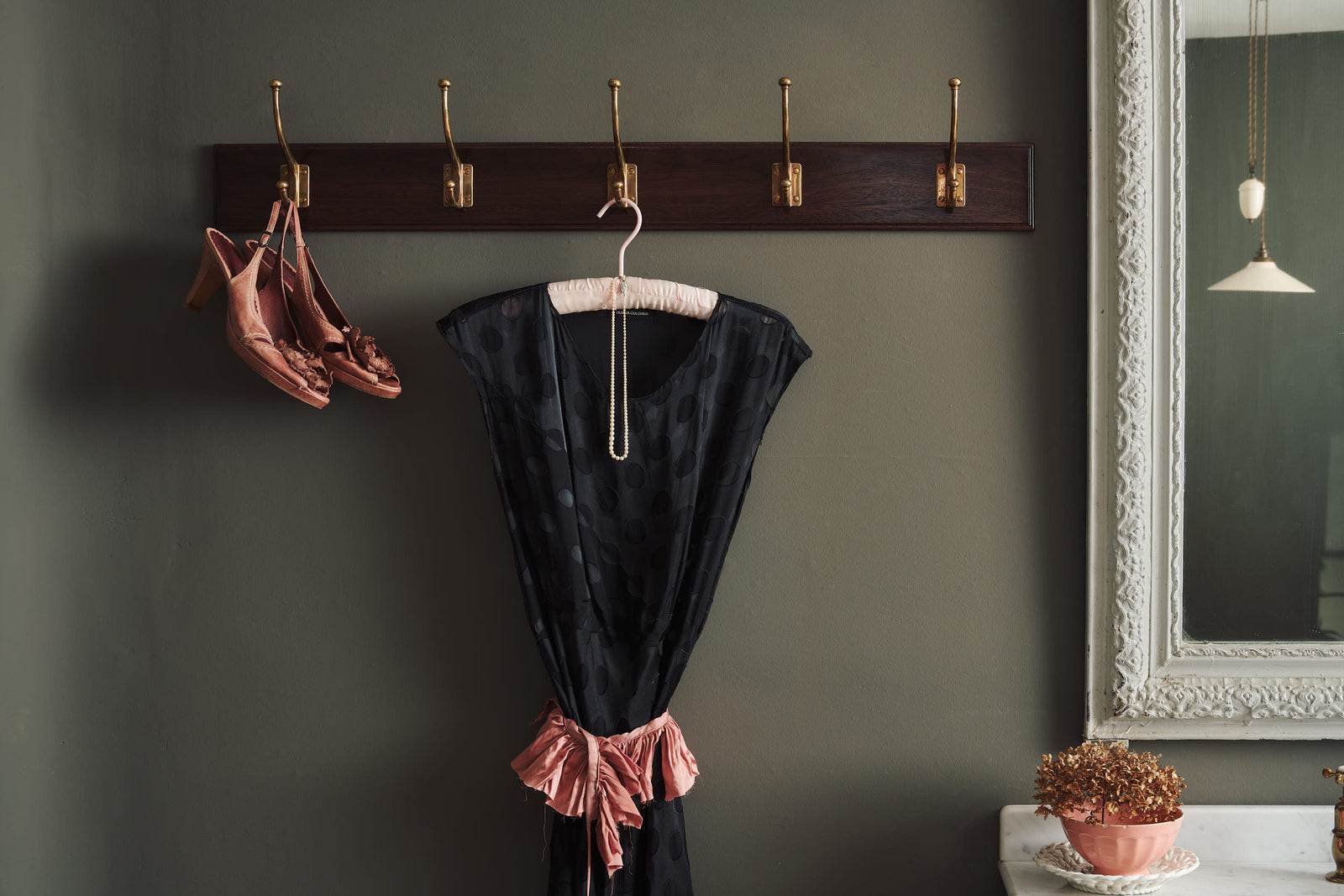 A Mahogany Finish Hook Rack with 5 Edwardian brass hooks, fitted to a moody grey wall with a black dress and dusky pink shoes hanging. A pink portrait painting and tall mirror decorate the space around the hook rack.