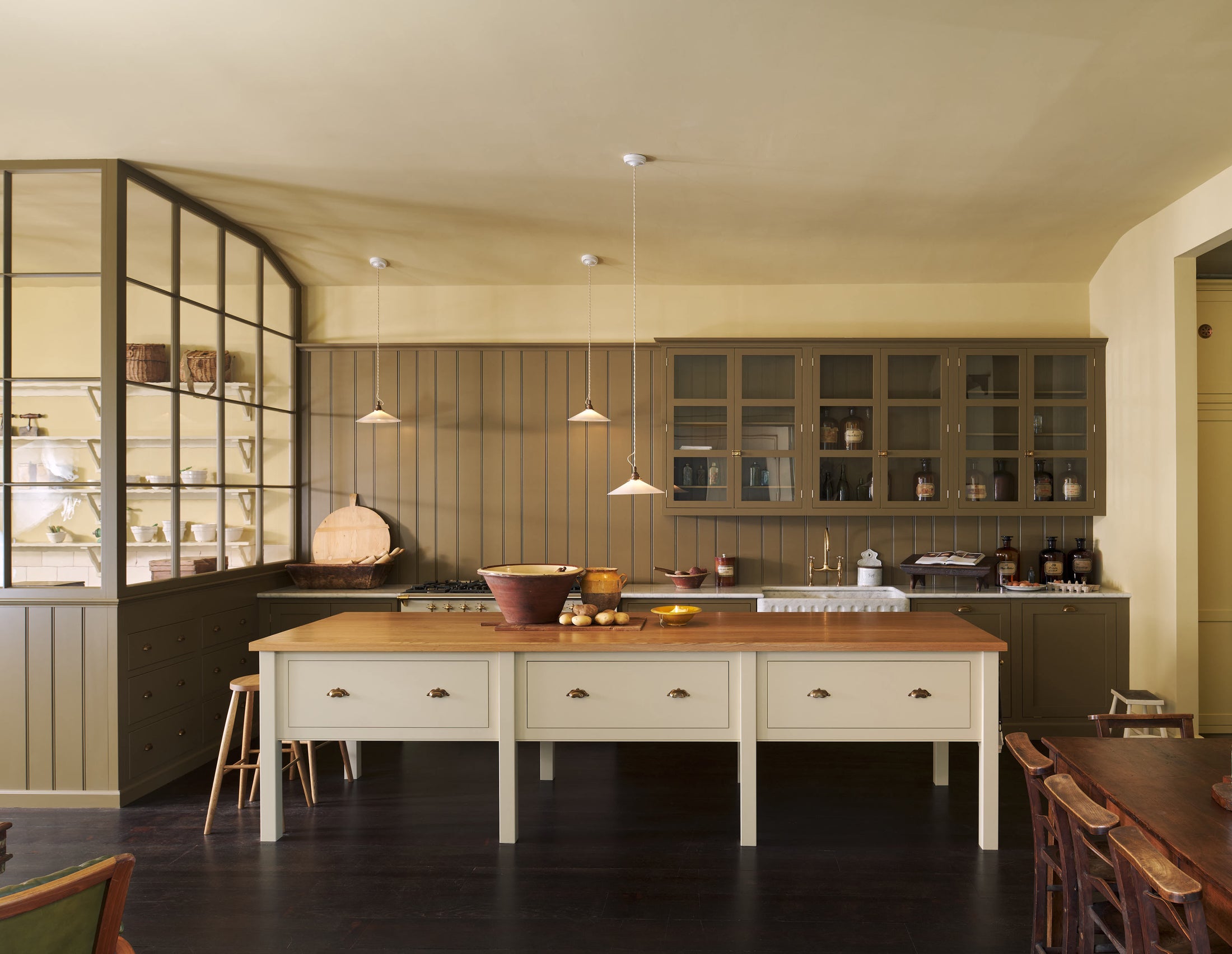 A simple Quaker cream and Folgate green Shaker kitchen features a cooker and sink run with Carrara marble worktop, glazed wall cupboards, and a bespoke prep table with a classic oak worktop. Lovingly adorned with milk pendant lights, artisan ceramics, antiques, and Helen stools, it creates a warm an