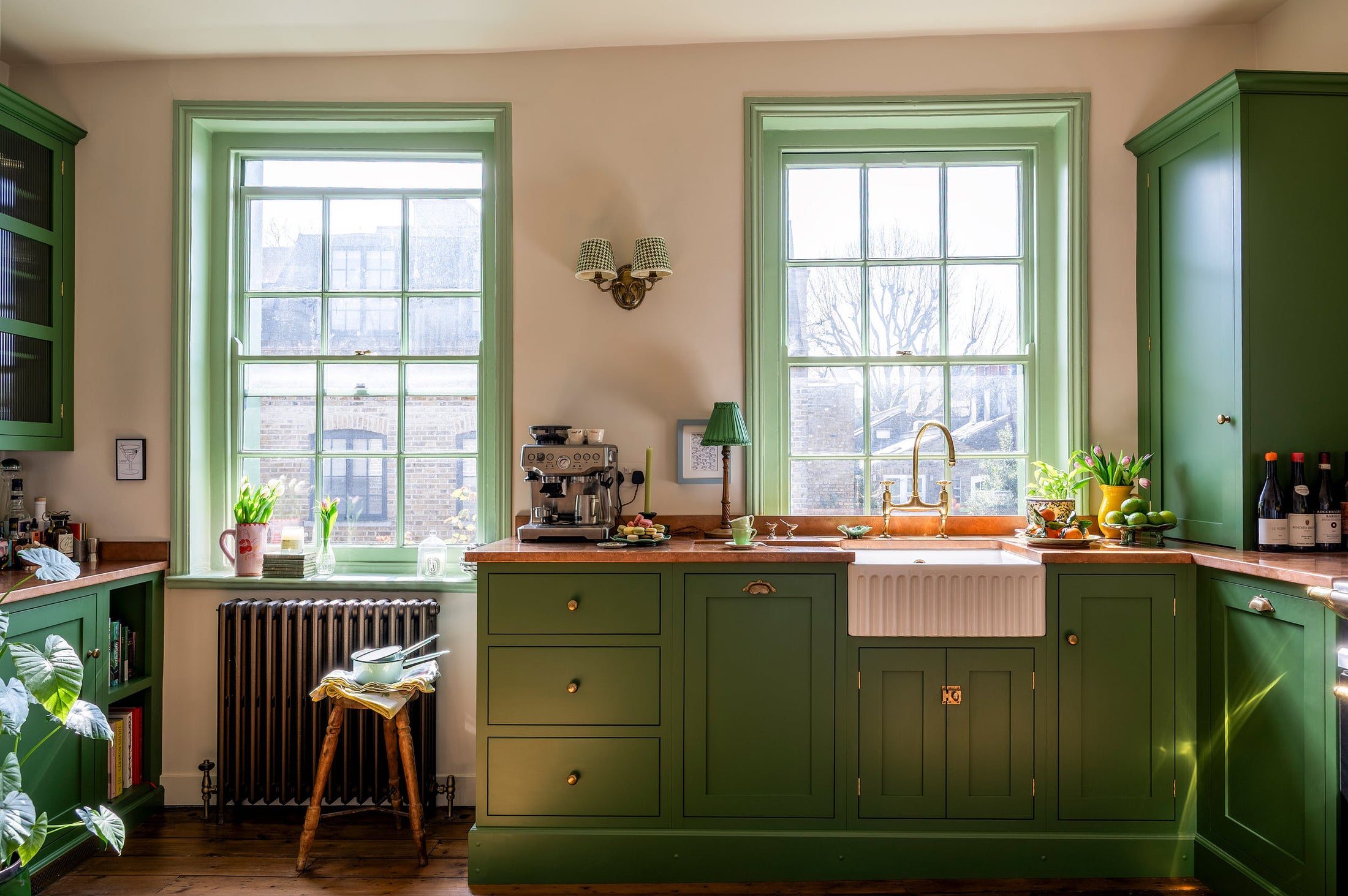 A bright and humble lime-green kitchen with an L-shaped cooker and sink run topped with aged copper worktops, featuring countertop and glazed cupboards, a single fluted Belfast sink with an aged brass Ionian tap, and colourful accents like tulips, a bowl of limes, macaroons, and assorted bottles of